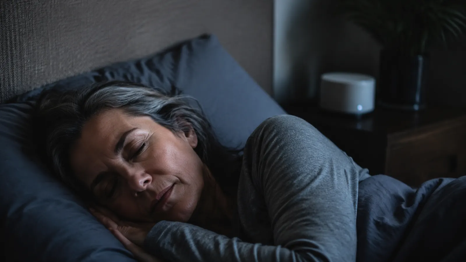 Woman sleeping peacefully in a dark bedroom with a white noise machine visible on the nightstand in the background